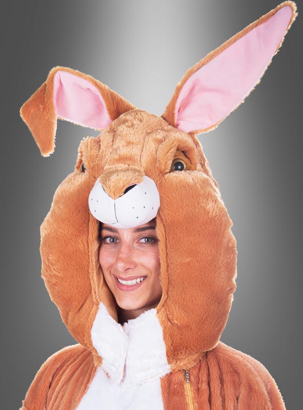 Eine Person, die lächelt, während sie das „Deluxe Hasenmaske Kopf Set waschbar“ trägt – ein braun-weißes Hasenkostüm mit großem Kopfteil, großen Ohren, rosa Futter und einem flauschigen Plüsch-Hasenkopf – vor einem grauen Hintergrund mit Farbverlauf.