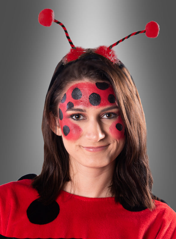 Eine Person in einem Marienkäferkostüm mit Gesichtsbemalung, einem rot-schwarz gepunkteten Käfer-Outfit und einem Stirnband mit Marienkäfer-Fühlern und roten Pompons vor einem grauen Hintergrund.