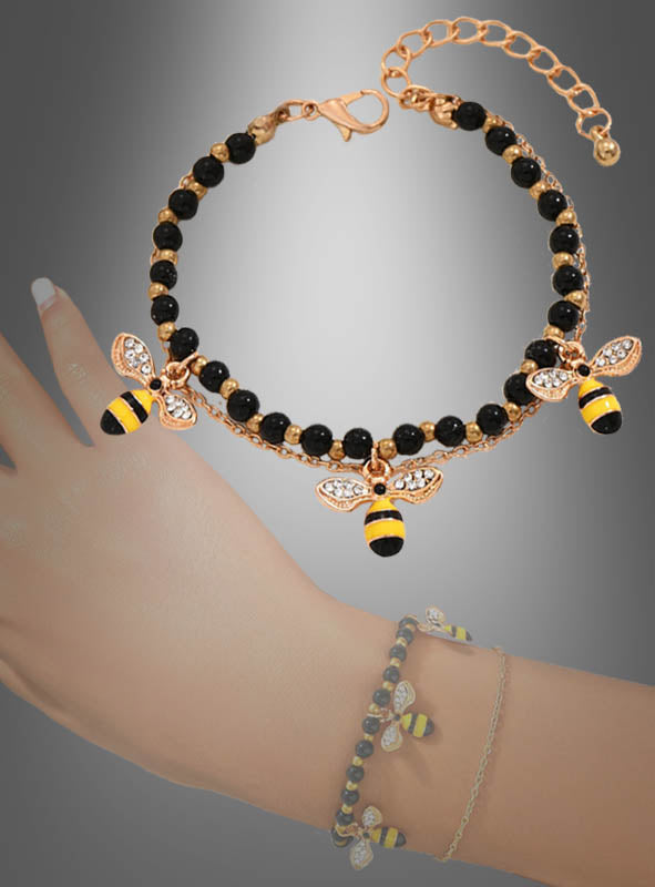 Der „Bienen Armband Kostümschmuck“ besteht aus einem goldenen Armband mit schwarzen Perlen und zwei Bienen-Anhängern, die Ihrem Look eine verspielte Note verleihen. Vor einem Farbverlaufshintergrund präsentiert und elegant am Handgelenk in Szene gesetzt, eignet sich dieses zarte Bettelarmband perfekt für jeden Anlass.