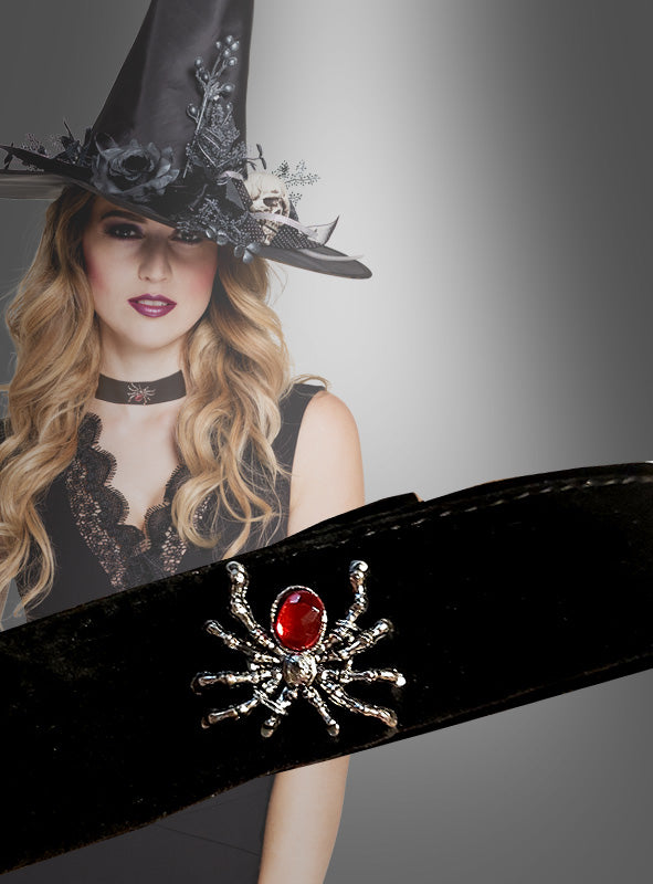 Eine Frau mit schwarzem Hexenhut und schwarzem Kleid trägt ein Samt-Halsband mit Spinne und Edelstein; dessen spinnenförmiger Anhänger mit einem roten Edelstein verleiht ihrem Outfit vor einem grauen Hintergrund mit Farbverlauf einen eleganten Hauch von Gothic-Schmuck.