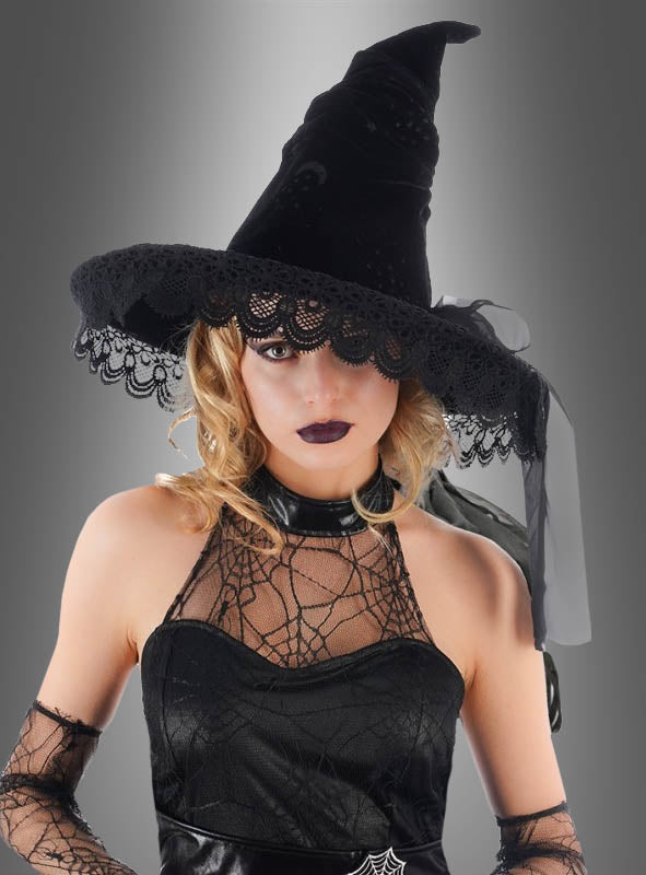 Eine Frau in einer edlen Hexenhütte aus Samt mit Spitze und einem Spinnennetzkleid, die dunklen Lippenstift trägt, posiert vor einem grauen Hintergrund – ein auffälliges Halloween-Kostüm, das perfekt zur Jahreszeit passt.