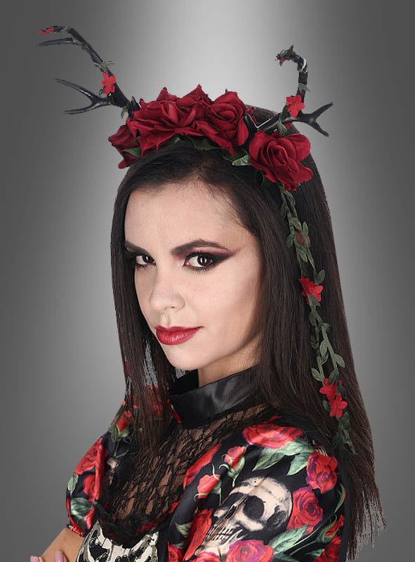 Eine Frau trägt ein Stirnband, das mit schwarzen Geweihen, dunkelroten Rosen und herabhängenden Ranken verziert ist – ein bezauberndes Beispiel für „Dark Mori“-Accessoires. Sie hat dunkles Haar, ein auffälliges Augen-Make-up und ein Kleid mit einem Totenkopf- und Rosenmuster, das perfekt zu ihrem „Deluxe Geweih-Haarschmuck mit Rosen“ passt.