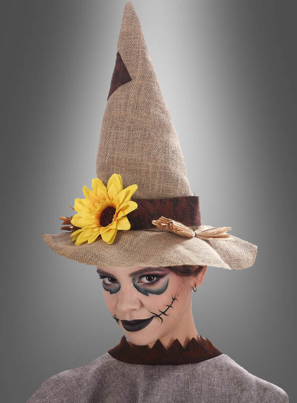 Eine Person in einem Vogelscheuchenkostüm aus Sackleinen mit dunklem Augen-Make-up und einem aufgenähten Mund, die vor einem grauen Hintergrund posiert und den „Hut Vogelscheuche mit Sonnenblume“ trägt – einen hohen Jutehut, der mit einer Sonnenblume verziert ist –, für das perfekte Halloween-Kostüm, das vom klassischen Vogelscheuchen-Look inspiriert ist.
