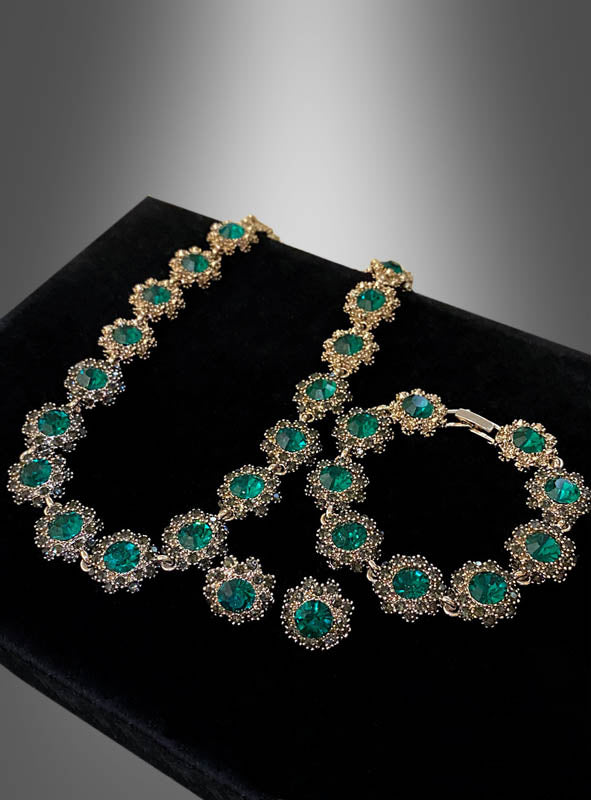 Das „Edles Schmuckset mit Strasssteinen, 4-teilig“ wird auf einer schwarzen Samtunterlage mit grau verlaufendem Hintergrund präsentiert und zeigt seine elegante goldene Halskette und das Armband, die mit großen grünen Edelsteinen und funkelnden Strasssteinen verziert sind – perfekt für jedes Kostüm im Stil der 20er Jahre.