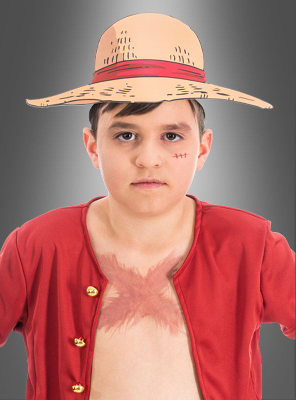 Ein Junge mit Strohhut, rotem, offenem Hemd und einer Narbe auf der Brust, verkleidet als Strohhut-Ruffy aus „One Piece“, steht vor einem grauen Hintergrund – die perfekte Inspiration für einen „One Piece“-Kindergeburtstag oder alle, die auf der Suche nach einzigartigen „One Piece“-Papierkopfbedeckungen sind.