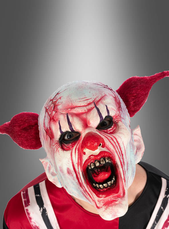 Eine Person, die die Horror-Clown-Maske aus Latex trägt, mit roten Ohren, einem weißen Gesicht, schwarzen Akzenten um die Augen, scharfen Zähnen und einem bedrohlichen roten Mund. Der graue Hintergrund mit Farbverlauf eignet sich perfekt für Ihre nächste Halloween-Party oder einen anderen gruseligen Anlass.