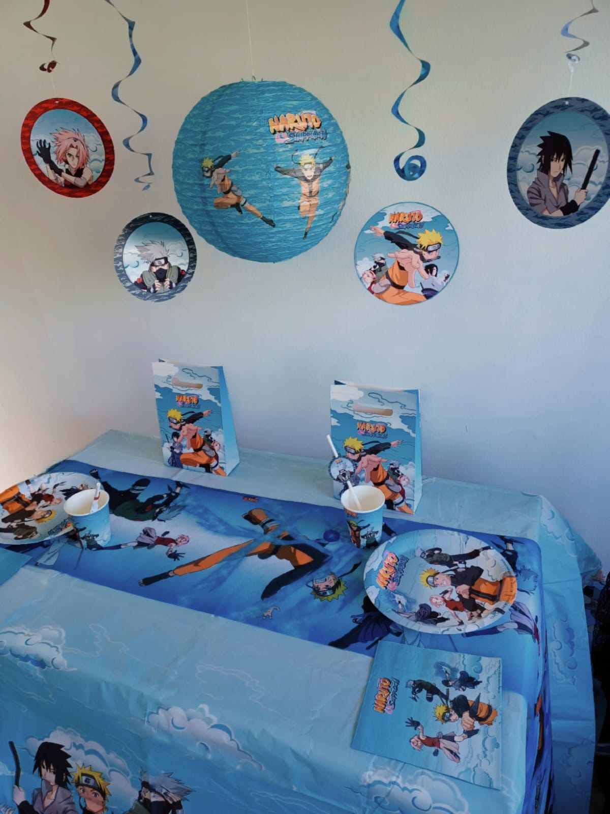 Ein gedeckter Tisch für eine Party zum Thema „Naruto“ mit Anime-Tischdekoration und einem farbenfrohen Tischläufer im Naruto-Manga-Stil sowie einer themenbezogenen Tischdecke, Tellern, Bechern, Tüten und Hängedekorationen, die Figuren aus dem Anime zeigen.