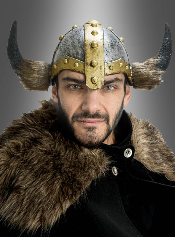 Eine Person, die den „Wikingerhelm Deluxe“ – einen Wikingerhelm aus Latex mit Hörnern – und einen mit Fell gefütterten Umhang trägt und mit neutralem Gesichtsausdruck direkt in die Kamera blickt, vor einem grauen Hintergrund.