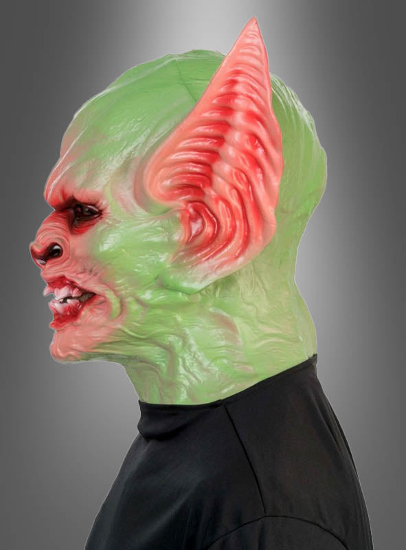 Eine Person, die die „Goblin Kobold Schaumstoff-Vollmaske Deluxe“ trägt – mit grüner und roter Farbgebung, übertrieben großen Ohren, Reißzähnen und faltiger Haut –, im Profil vor einem grau verlaufenden Hintergrund.