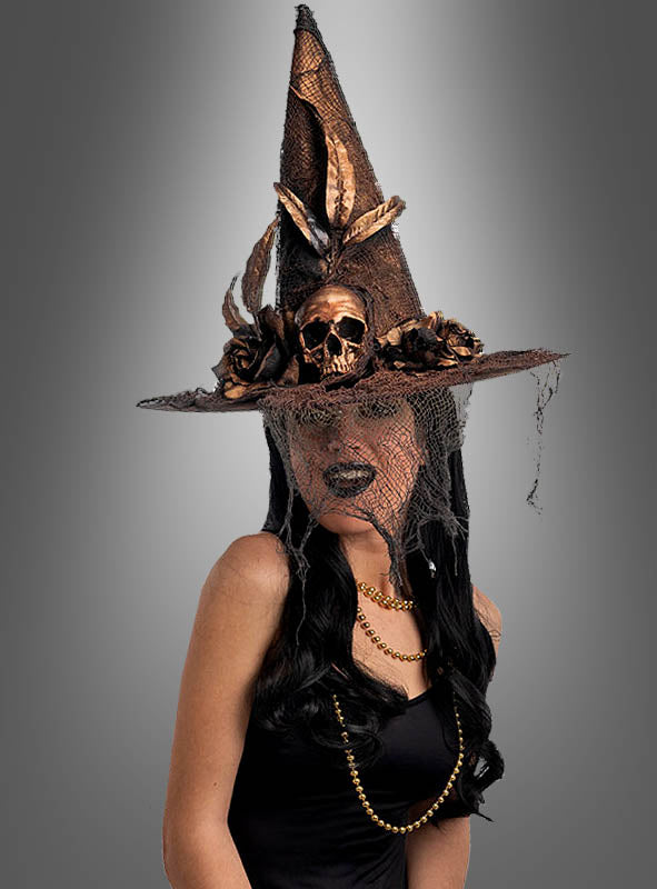 Eine Frau in einem schwarzen Kleid und mit einer Halskette trägt den hohen, spitzen Hexenhut mit Totenkopf in Kupfer-Gold, der mit Blumen, Federn und einem Netzschleier verziert ist und vor einem grauen Hintergrund einen auffälligen Hexenhut im Shabby-Look-Stil bildet.