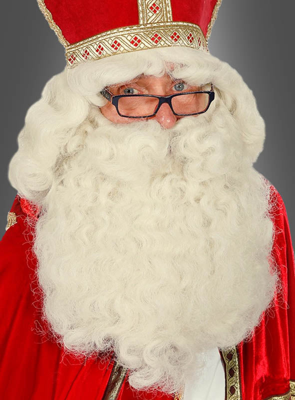 Eine Person, die eine rot-goldene Robe und eine Mitra trägt, mit der „Deluxe Weihnachtsmann-Perücke und Bart“ – einem Set aus einer großen, weißen, lockigen Perücke und einem Bart – sowie einer Brille, blickt vor einem grauen Hintergrund in die Kamera.