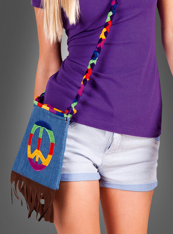 Eine Person, die ein lila Shirt und helle Jeansshorts trägt, hat die „Hippie Jeans Handtasche“ dabei – eine kleine Jeanshandtasche mit Fransen, einem Regenbogen-Friedenssymbol und einem bunten, geflochtenen Riemen, die sich perfekt als Accessoire für ein Hippie-Kostüm eignet.