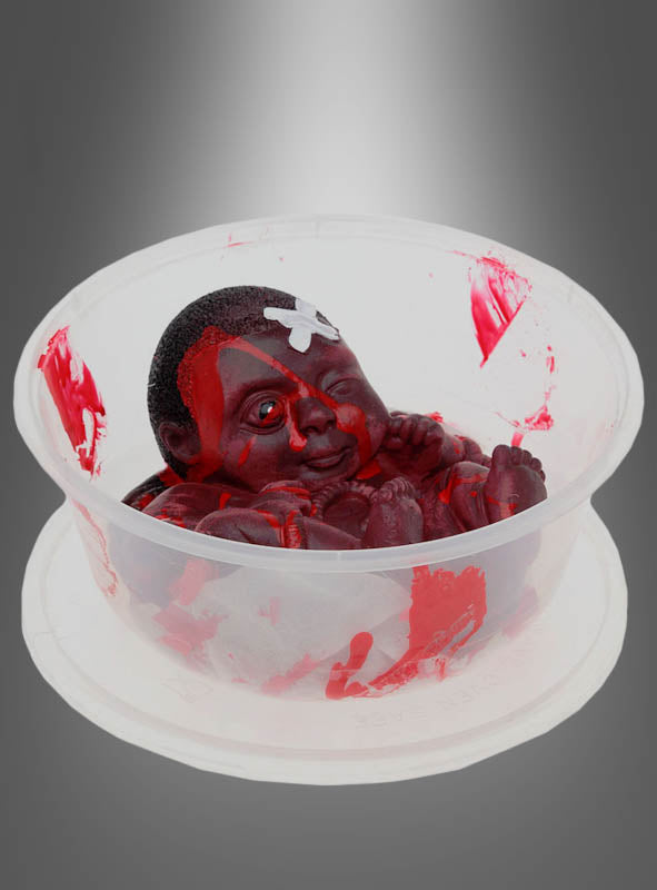 In einem Plastikbehälter befindet sich das „Blutiges Horror Baby in Schüssel“ – eine kleine, dunkelhäutige Babypuppe, die mit roter Farbe bespritzt ist und auf der Stirn einen weißen Fleck aufweist, was einen gruseligen Effekt erzeugt. Vor einem grauen Hintergrund mit Farbverlauf ist dieses Stück die perfekte Zombie-Dekoration und verleiht jeder Halloween-Dekoration eine unheimliche Note.