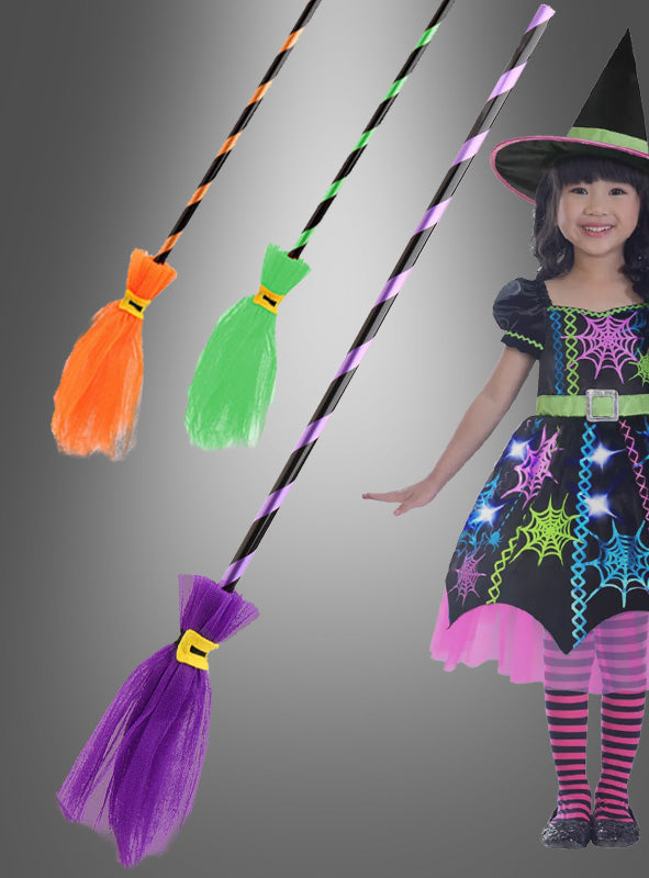 Drei Hexenbesen mit Tüll (80 cm), jeweils mit leuchtenden Streifen in Orange, Grün und Lila – perfekt als Hexenbesen für Kinder – liegen neben einem Kind, das ein buntes Hexenkostüm mit Hut, Kleid und gestreiften Strumpfhosen trägt. Diese farbenfrohen Hexenbesen mit Tüll (80 cm) sind das ideale Hexenkostüm-Accessoire, um fantasievolles Spielen anzuregen.