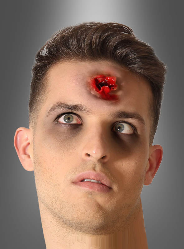 Ein Mann mit dunklen Ringen unter den Augen trägt eine große, blutige Latexnarbe in Form einer Schusswunde mitten auf der Stirn, was sein Zombie-Kostüm noch furchterregender macht.