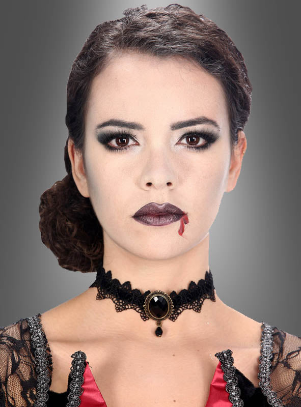 Eine Frau mit blassem Make-up, dunklem Lidschatten und Lippenstift trägt ein schwarzes Spitzenhalsband im Gothic-Stil und ein rotes Kostüm; auf ihrer Lippe ist Kunstblut zu sehen, sodass sie vor einem grauen Hintergrund wie ein Vampir wirkt.