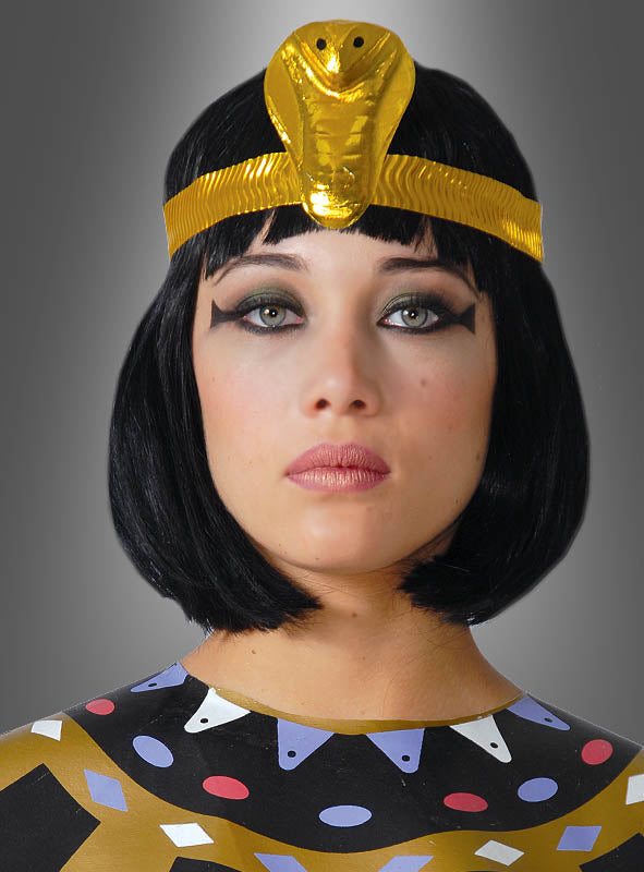 Eine Person mit einer schwarzen Bob-Perücke, dem „Goldene Kobra“-Kopfschmuck für Kleopatra und einem Outfit mit geometrischen Mustern sowie auffälligem schwarzem Eyeliner, die vor einem grauen Hintergrund mit Farbverlauf posiert.