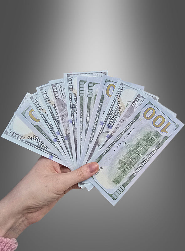 Eine Hand hält einen aufgefächerten Stapel von sieben 100-Dollar-Scheinen mit der Aufschrift „Scheine Spielgeld für Millionäre“, die wie hochwertige gefälschte Dollarscheine aussehen, vor einem grauen Hintergrund mit Farbverlauf.