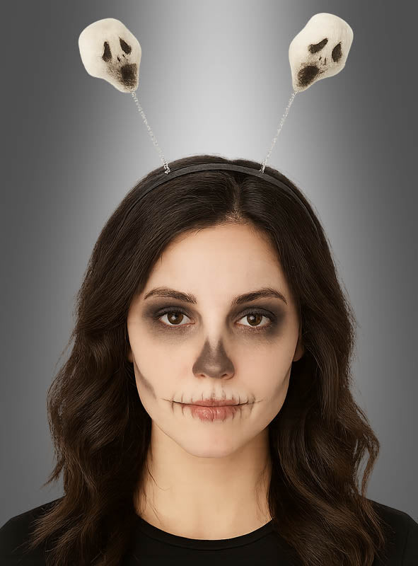 Eine Frau mit dunklem, schulterlangem Haar trägt Gesichtsbemalung mit Totenkopfmotiven und einen Halloween-Haarreif mit Geistern, der mit zwei kleinen Totenköpfen an Federn verziert ist, vor einem grauen Hintergrund.