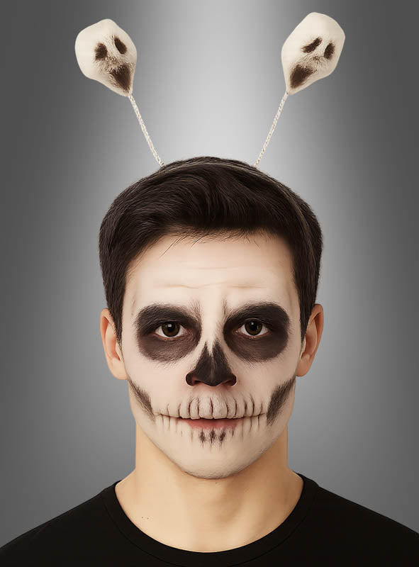 Eine Person mit einer Gesichtsbemalung, die einem Schädel ähnelt, und einem Halloween-Haarreif mit Geistern – einem Stirnband mit zwei auf Federn befestigten Geistern aus Schaumstoff – vor einem neutralen Hintergrund mit Farbverlauf.