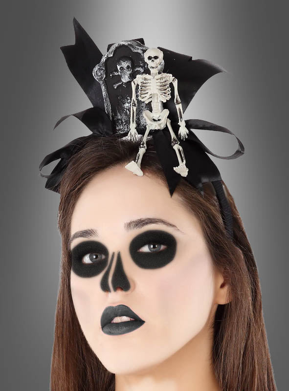 Eine Person mit dunklem, schädelartigem Make-up trägt einen „Skelett Haarreif Halloween“, der mit einem kleinen Skelett und einem Grabstein verziert ist, vor einem grauen Hintergrund mit Farbverlauf.