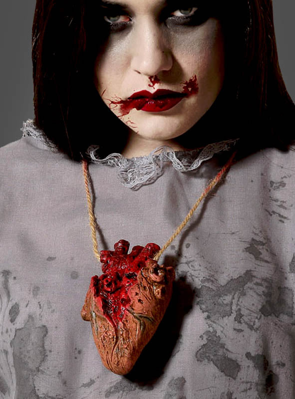 Eine Person mit auffälligem Make-up und blutroten Lippen trägt die Halloween-Halskette „Blutiges Herz Deko“ – eine Halskette mit einem realistisch gestalteten, blutigen Herz an einer Kordel – über einem grauen, fleckigen Shirt, was eine gewagte Wahl an Halloween-Schmuck darstellt.