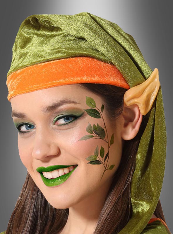 Eine Person mit einer grün-orangefarbenen Elfenmütze, Latex-Ohrspitzen für Elfen, grünem Make-up, grünem Lippenstift und einem Gesichts-Make-up in Form einer Blattpflanze lächelt vor einem grauen Hintergrund mit Farbverlauf in die Kamera.