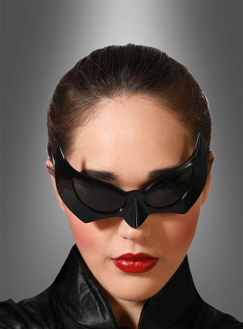 Eine Person mit nach hinten gebundenen dunklen Haaren, die die „Fledermaus-Brille“-Halloween-Katzenaugenmaske, roten Lippenstift und ein schwarzes Outfit trägt – ein elegantes Kostümaccessoire für Halloween oder Karneval –, blickt vor einem grauen Hintergrund mit Farbverlauf nach vorne.