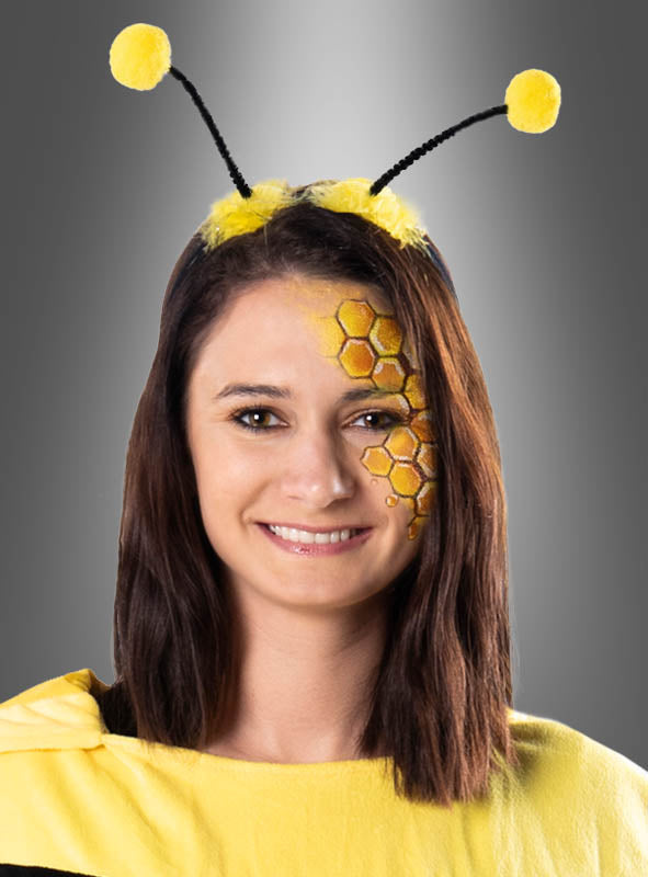 Eine Frau in einem Bienenkostüm mit einem Haarreif in Form von Bienenantennen mit gelben Pompons und einer Waben-Gesichtsbemalung auf einer Wange lächelt vor einem grauen Hintergrund in die Kamera.