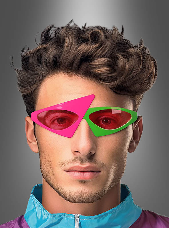 Ein Mann mit kurzen braunen Haaren trägt eine asymmetrische 80er-Jahre-Brille – zwei ungleiche geometrische Sonnenbrillengläser, eines in Rosa und eines in Grün – sowie eine lila-blaue Jacke vor einem grauen Hintergrund mit Farbverlauf.