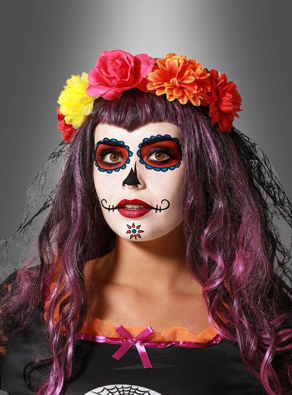 Eine Person mit einem Make-up im Stil des „Tages der Toten“ (Dia de los Muertos), die eine lila Perücke trägt und das bunte „Sugar Skull Blumen“-Haarband, das mit roten, gelben und orangefarbenen Blumen verziert ist, steht vor einem grauen Hintergrund.