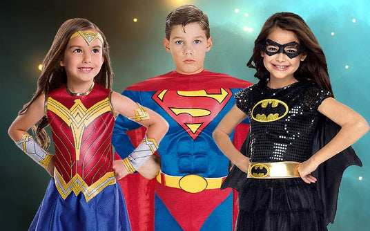 Drei Kinder tragen Superheldenkostüme: Wonder Woman, Superman und Batgirl. Sie posieren mit den Händen in den Hüften vor einem blauen und dunklen Hintergrund mit hellen Flecken.