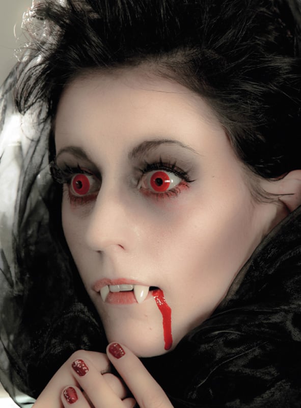 Eine Person mit blasser Haut, roten Augen, die durch die Jahreslinsen „Red Devil Rot“ noch stärker hervorgehoben werden, Vampirzähnen und Kunstblut auf den Lippen; sie trägt ein schwarzes Kostüm und dunkles Make-up und blickt zur Seite.