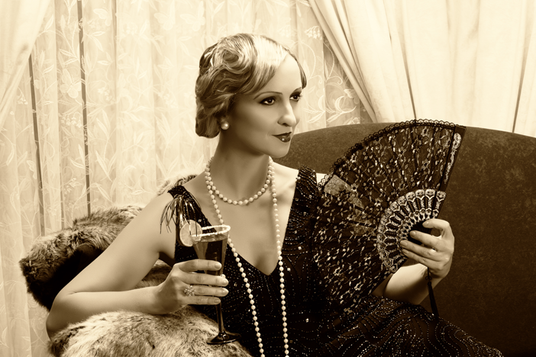 Eine Frau in Kleidung aus den 1920er Jahren hält einen mit Perlen verzierten Fächer und einen Cocktail in der Hand; sie trägt eine Perlenkette und ein Flapper-Kleid und sitzt auf einem Pelzüberwurf vor einem Vorhang als Hintergrund. Sepiafarbene Aufnahme.