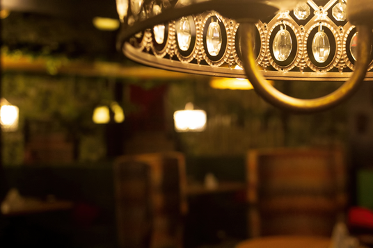 Im Vordergrund eines schummrig beleuchteten Restaurants hängt ein Kronleuchter mit Kristallverzierungen, während im Hintergrund gepolsterte Stühle und Tische verschwommen zu sehen sind.
