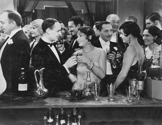 Eine überfüllte, formelle Partyszene aus dem frühen 20. Jahrhundert zeigt Männer in Smokings und Frauen in Abendkleidern, die an einer Bar miteinander plaudern und ihre Getränke in der Hand halten.