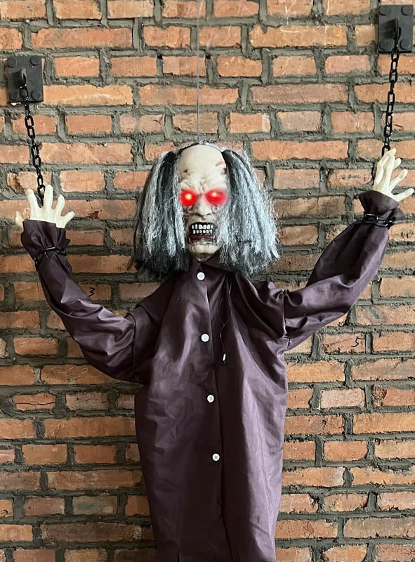 Wir präsentieren den „Lebensgroßen Zombie 185 cm an Ketten animiert“ – eine furchterregende Halloween-Dekoration mit leuchtend roten Augen, verfilztem grauen Haar und Ketten an den Handgelenken, die an einer Backsteinwand befestigt ist.