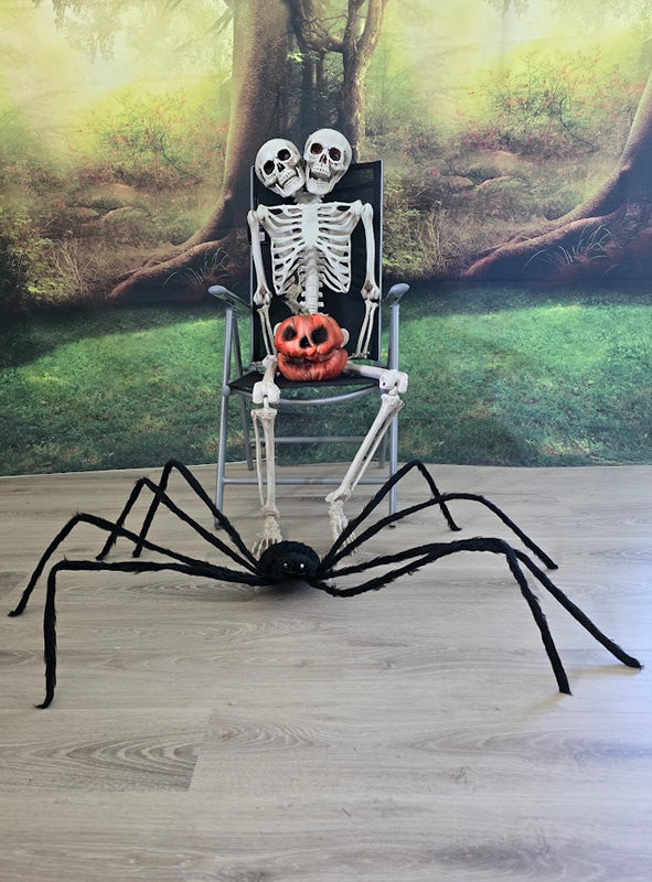 Zwei Skelette sitzen auf einem Stuhl, auf ihrem Schoß eine Kürbislaterne, während sich vor ihnen eine 150 cm große schwarze Riesenspinne mit LED-Augen auf dem Boden ausbreitet. Im Hintergrund ist ein gruseliges Waldwandbild zu sehen, das diese unheimliche Halloween-Spinnendekoration abrundet.