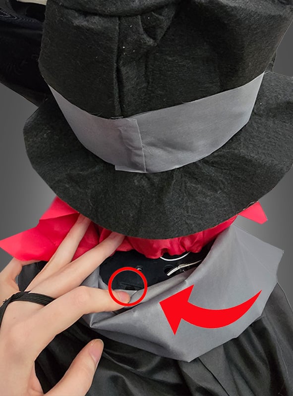 Eine Hand rückt den Kragen des „Friedhof Skelett 38 cm mit Animation“ zurecht, das einen großen Hut trägt, der an ein klassisches animiertes Skelett erinnert; ein roter Pfeil weist auf ein kleines schwarzes Mikrofon hin, das unauffällig im Kragen befestigt ist – ideal, um Ihre Halloween-Dekoration aufzuwerten.