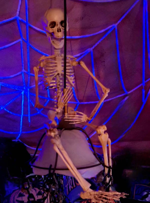Eine Skelett-Dekoration aus Kunststoff thront auf einer Leuchte, vor dem Hintergrund des „Riesigen UV-Spinnennetzes 300 cm, weiß“ – so entsteht eine gruselige Halloween-Dekoration, die durch violette Beleuchtung und weiße Spinnennetz-Details noch verstärkt wird.
