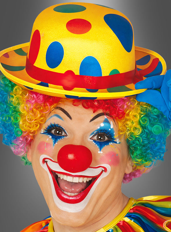 Eine als Clown verkleidete Person mit einer Regenbogenperücke, einem bunten Clownhut mit Punkten, einer roten Nase und übertriebenem Make-up lächelt breit vor einem grauen Hintergrund.