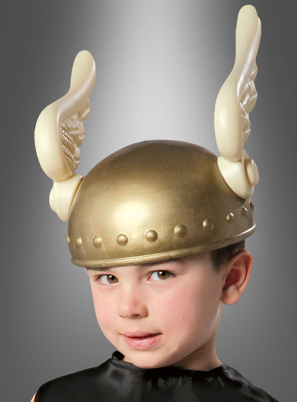 Ein kleines Kind trägt einen goldenen „Kleiner Wikinger Helm Kinder“-Helm mit großen, flügelförmigen Verzierungen auf beiden Seiten vor einem grauen Hintergrund mit Farbverlauf.