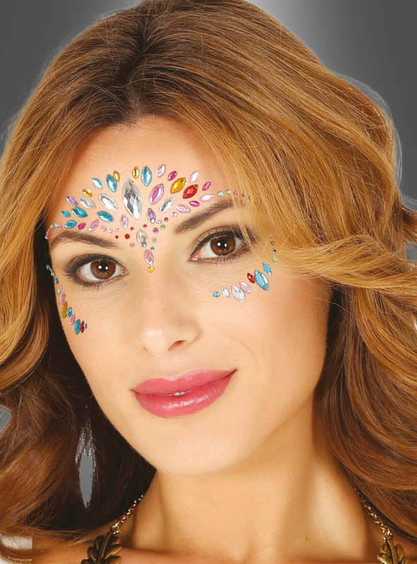Eine Frau mit welligem hellbraunem Haar und glänzendem rosa Lippenstift, die bunte selbstklebende Strasssteine für das Gesicht trägt, die symmetrisch auf ihrer Stirn und um ihre Augen herum angeordnet sind, vor einem grauen Hintergrund.