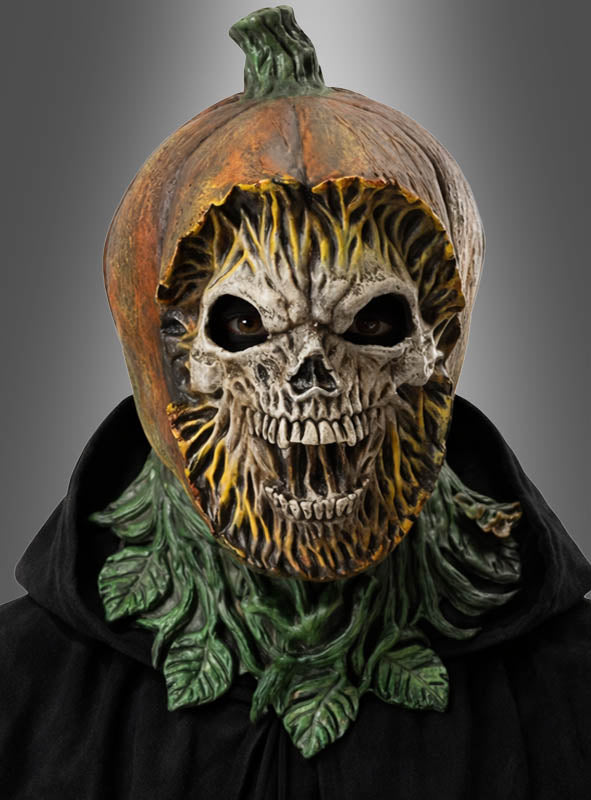 Eine Person trägt die „Gruselige Kürbis-Maske aus Latex“ mit einem Totenkopfgesicht, dunklen Augenlöchern und grünen Blattakzenten, dazu einen schwarzen Umhang vor einem grauen Hintergrund mit Farbverlauf. Diese auffällige Halloween-Maske sorgt für einen unheimlichen und zugleich festlichen Auftritt.