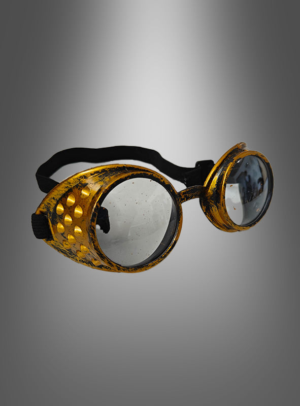 Ein Paar goldene Steampunk-Schweißerbrillen mit runden, klaren Gläsern im Vintage-Stil, einem schwarzen Riemen und strukturierten Seitenteilen – perfekt als Steampunk-Brille oder als Blickfang für Ihr Steampunk-Kostüm – präsentiert vor einem grauen Hintergrund mit Farbverlauf.
