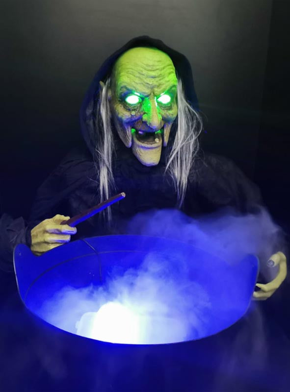 Eine Hexe mit grünem Gesicht, leuchtenden Augen und einem Zauberstab steht über einem Kessel, aus dem blaues Licht und Nebel strömen, wie bei einer „Bunte LED Mini Nebelmaschine“.