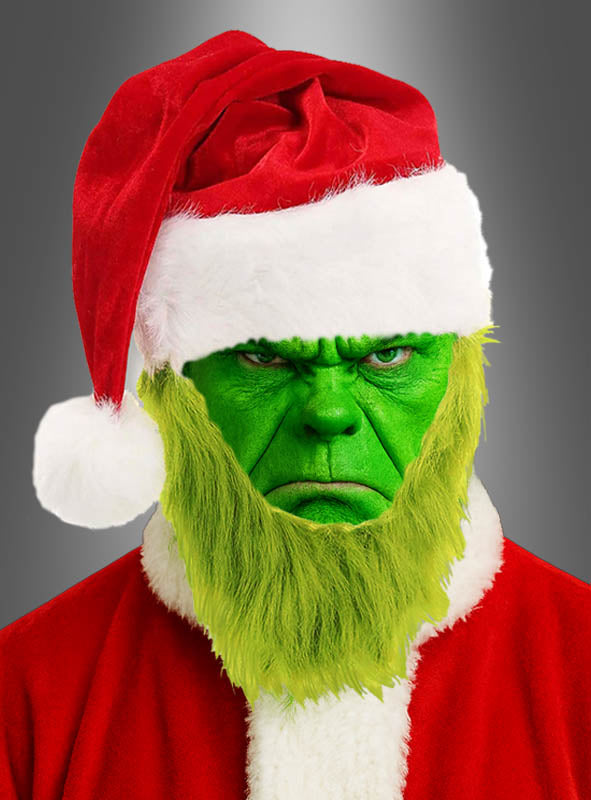 Eine Person mit grünem Gesicht und gelbgrünem Bart trägt eine Nikolausmütze mit grünem Bart und ein Weihnachtsmannkostüm, das dem Grinch-Kostüm ähnelt, vor einem schlichten grauen Hintergrund.