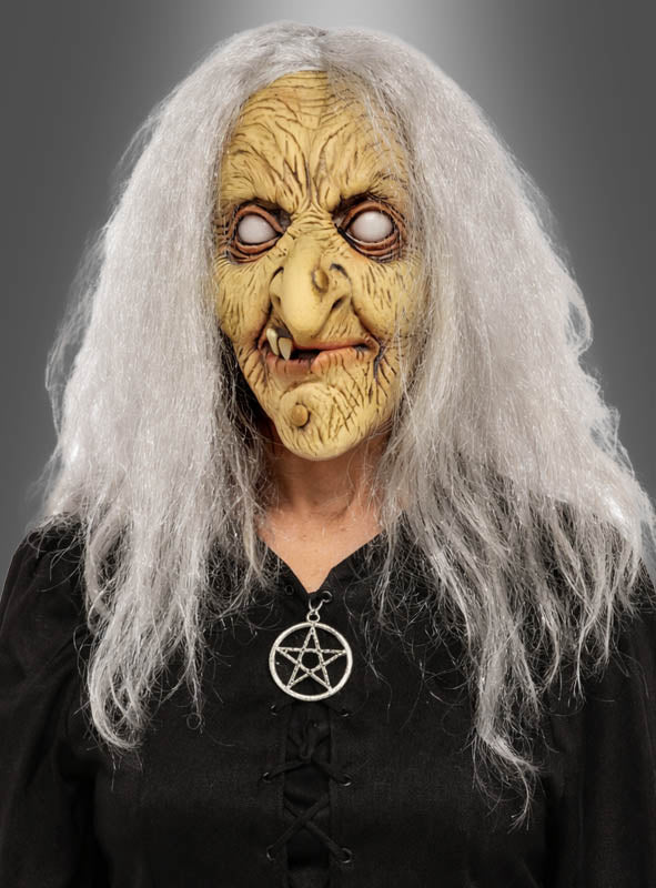 Sie trägt die „Hexen Maske Latex mit Haaren“ mit weißen Haaren, ist in schwarze Kleidung gekleidet und trägt eine Pentagramm-Halskette, vor einem grauen Hintergrund.