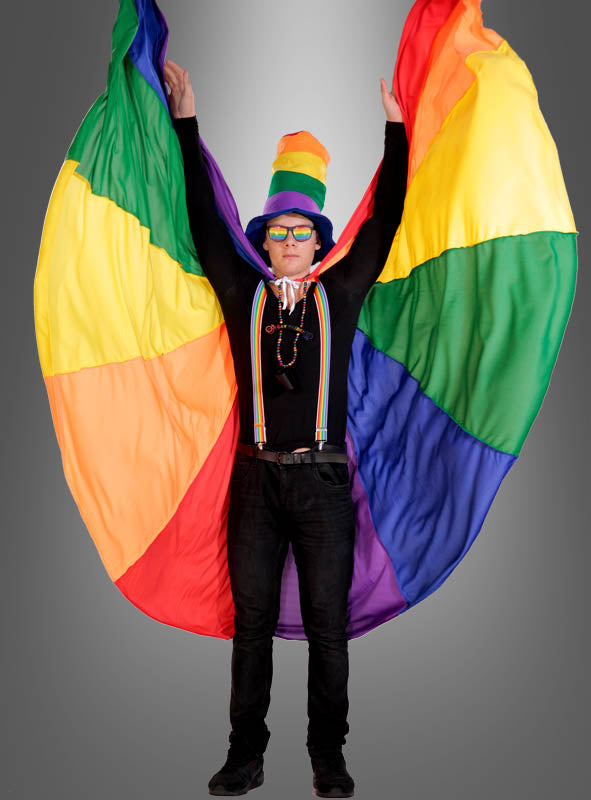 Eine Person, die einen Regenbogenhut, eine Sonnenbrille und Hosenträger trägt, hält den „Deluxe Umhang Cape Regenbogen 7 Meter“ hinter sich wie einen bunten Umhang hoch und erzeugt so einen auffälligen, flügelartigen Effekt. Dieser farbenfrohe Umhang eignet sich perfekt als Karnevalskostüm oder Umhang für Damen und Herren und hebt sich eindrucksvoll vom schlichten grauen Hintergrund ab.