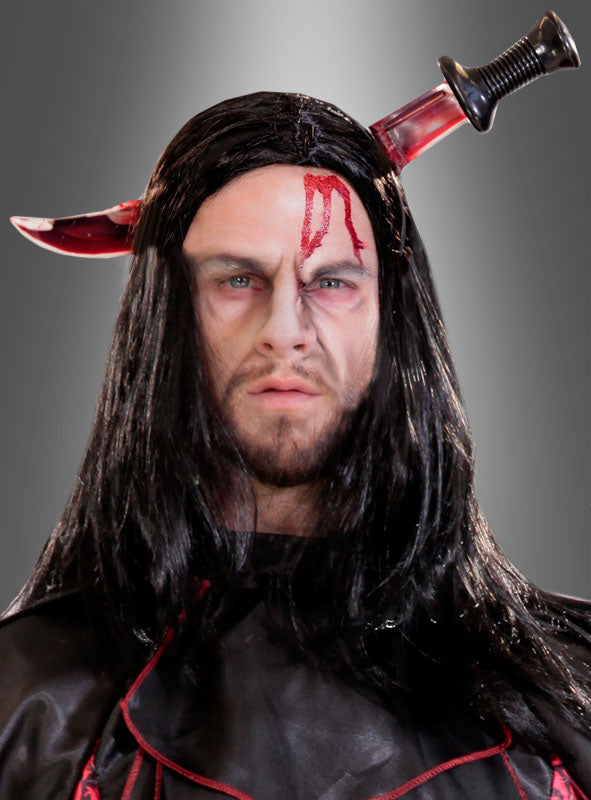 Eine Person, die eine schwarze Perücke und ein Kostüm trägt, ergänzt durch die Attrappe „Deluxe blutendes Messer im Kopf“ – ein Horror-Halloween-Accessoire, das den Anschein erweckt, als stecke ein blutiges Messer in ihrem Kopf.